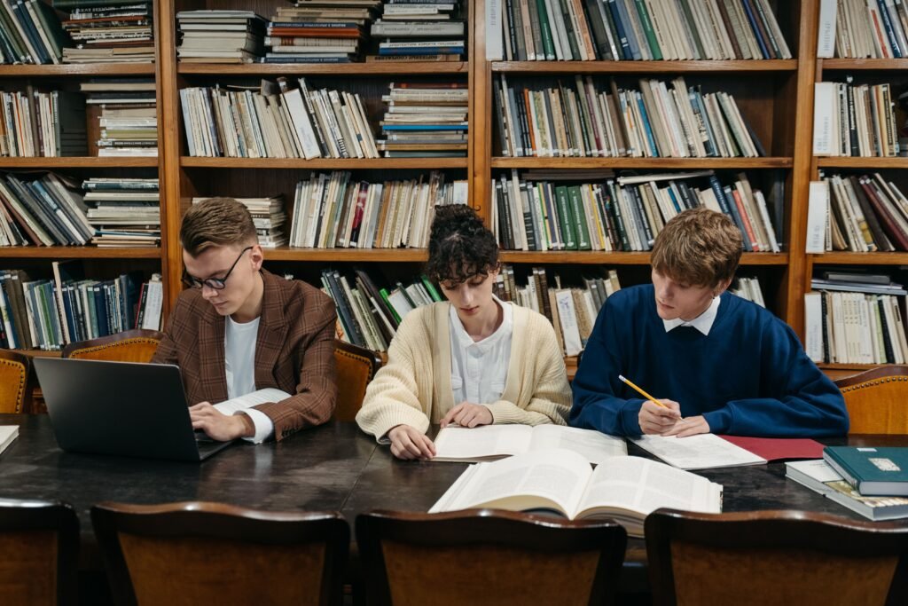 Three students work on laptops and books in a university library filled with shelves.